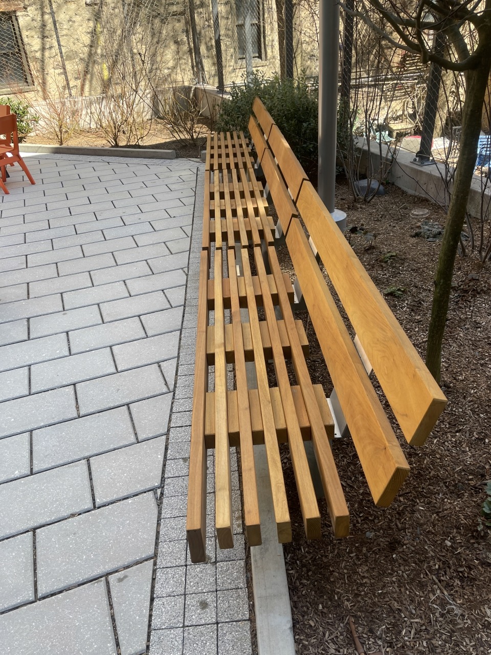 Long, modern wooden bench situated in a landscaped courtyard with paved stone pathway, providing a tranquil seating area for relaxation.